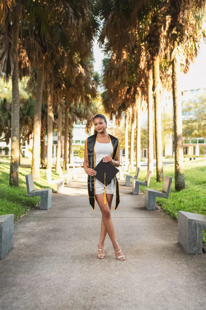 A UCF graduate in a white dress holding her cap while standing on a concrete walkway lined with tall palm trees on the UCF campus.