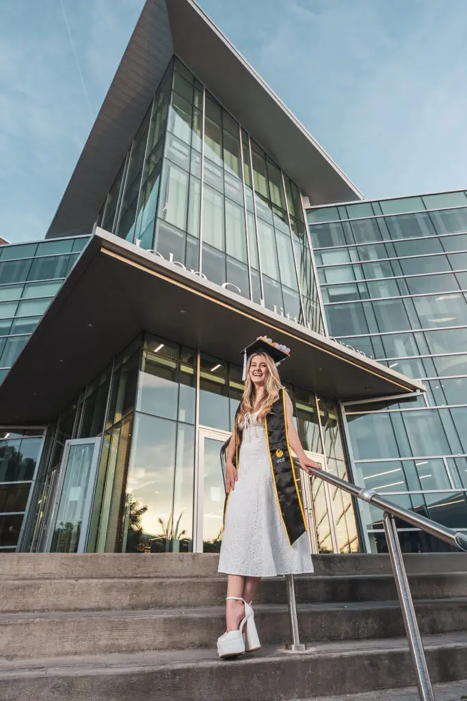 A smiling UCF graduate in a white dress and a black and gold UCF stole, posing on the steps of the John C. Hitt Library for a professional graduation photo.
