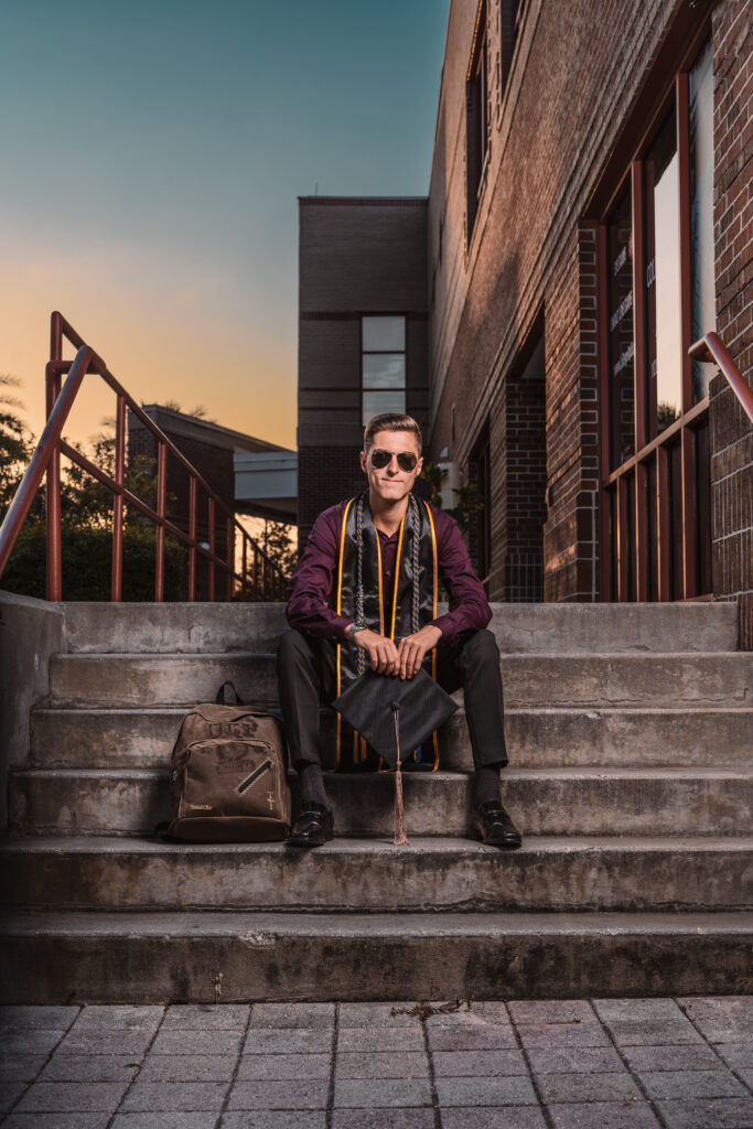 A male UCF graduate wearing sunglasses and a maroon shirt sitting on concrete steps at sunset, holding his cap next to a vintage-style UCF backpack.