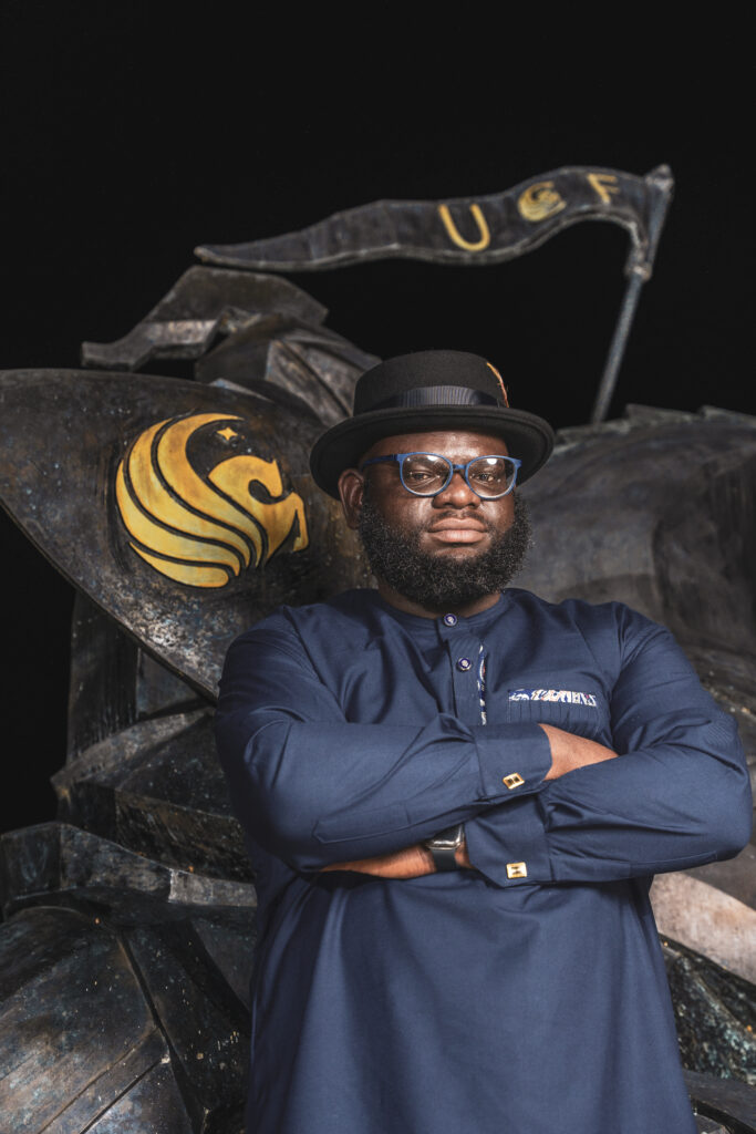 A UCF senior posing with arms crossed in front of the Charging Knight statue at night, wearing a fedora and blue formal attire during a dramatic graduation photoshoot.